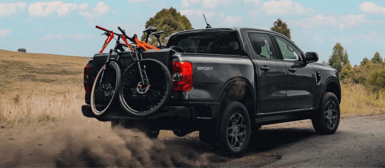 A dark grey pickup truck carrying mountain bikes kicks up dust on a dirt road against a backdrop of rolling hills and a cloudy sky.
