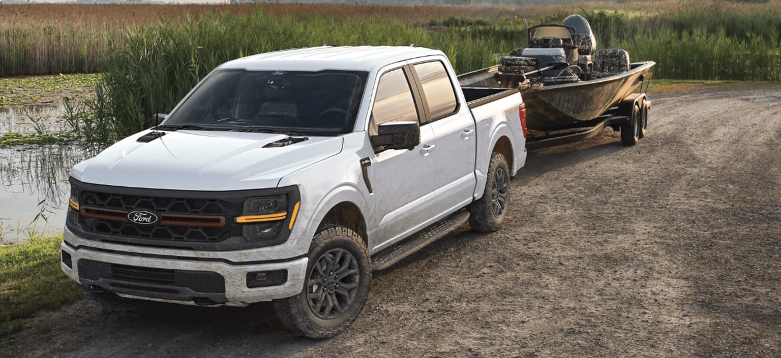 White pickup truck with boat trailer parked on dirt road near body of water.