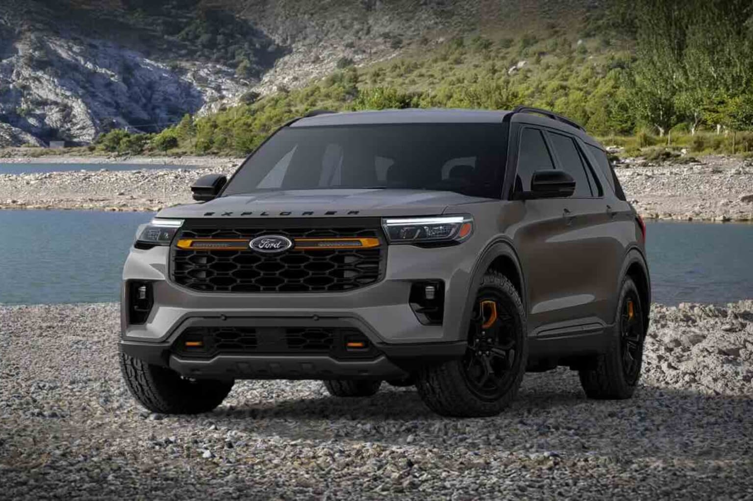 A gray SUV sits on a rocky shore with a calm body of water and a mountainous landscape in the background.