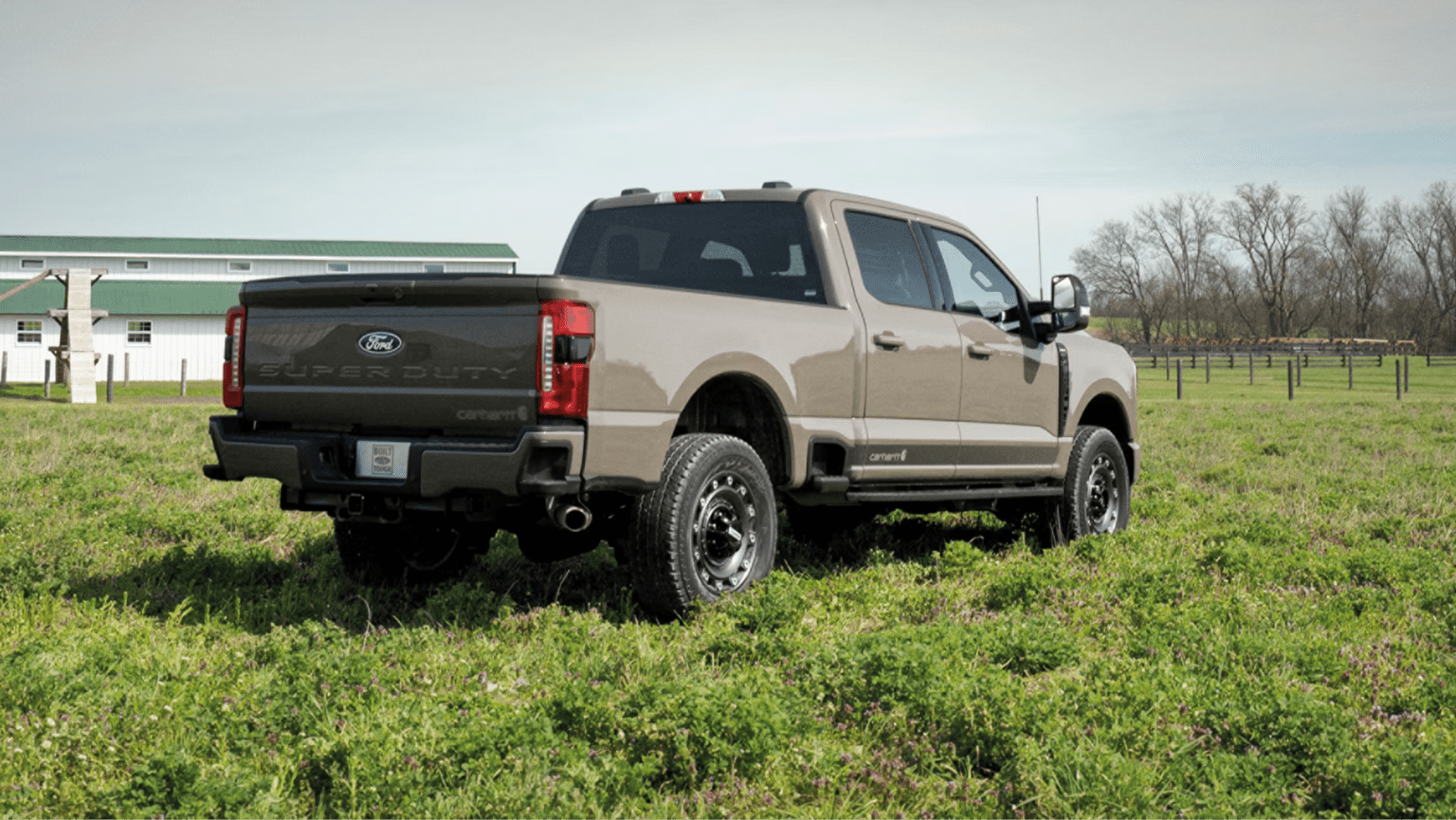 A tan pickup truck in a field with a white barn and trees in the background.