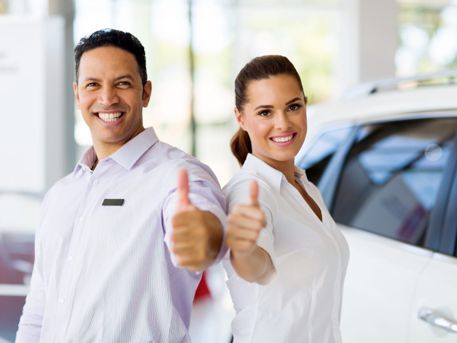 Two smiling individuals, a man and a woman, are standing in front of a car dealership, giving a thumbs up gesture.
