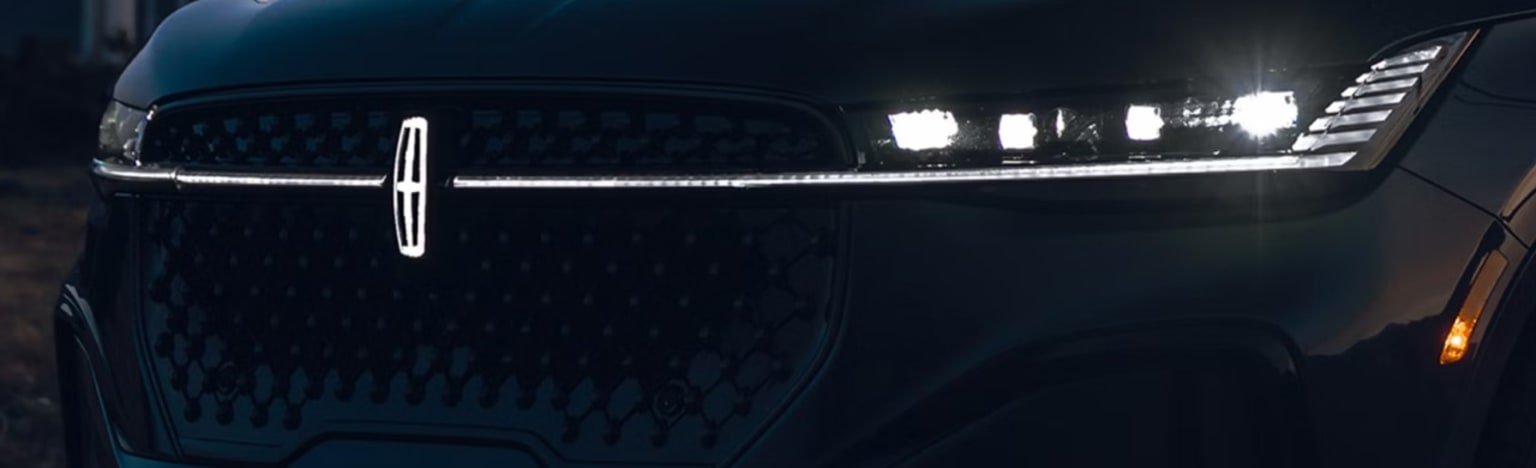 A dark car grille with illuminated headlights is set against a blurred, dark background.