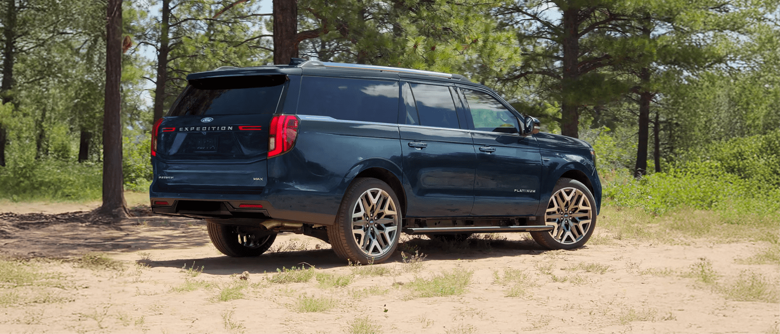 A large dark blue SUV parked on a sandy clearing surrounded by a dense forest of pine trees.