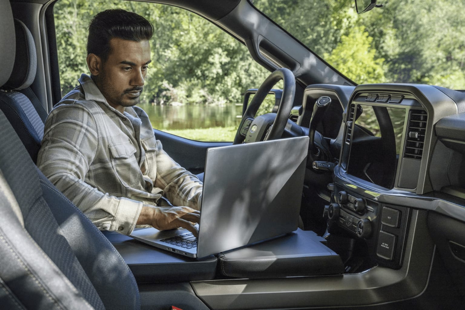 A person sits in the driver's seat of a vehicle, intently focused on a laptop computer placed on their lap as the lush, green landscape outside the car window provides a scenic backdrop.