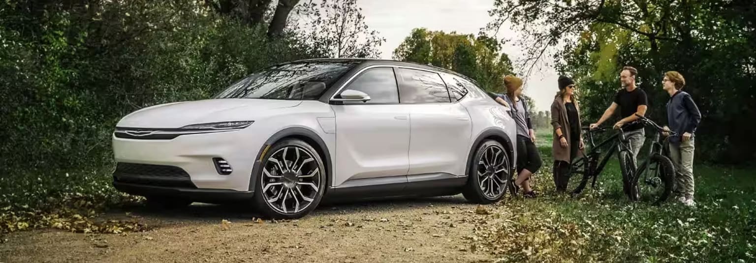 a couple of people standing next to a white car on a dirt road next to a tree filled field