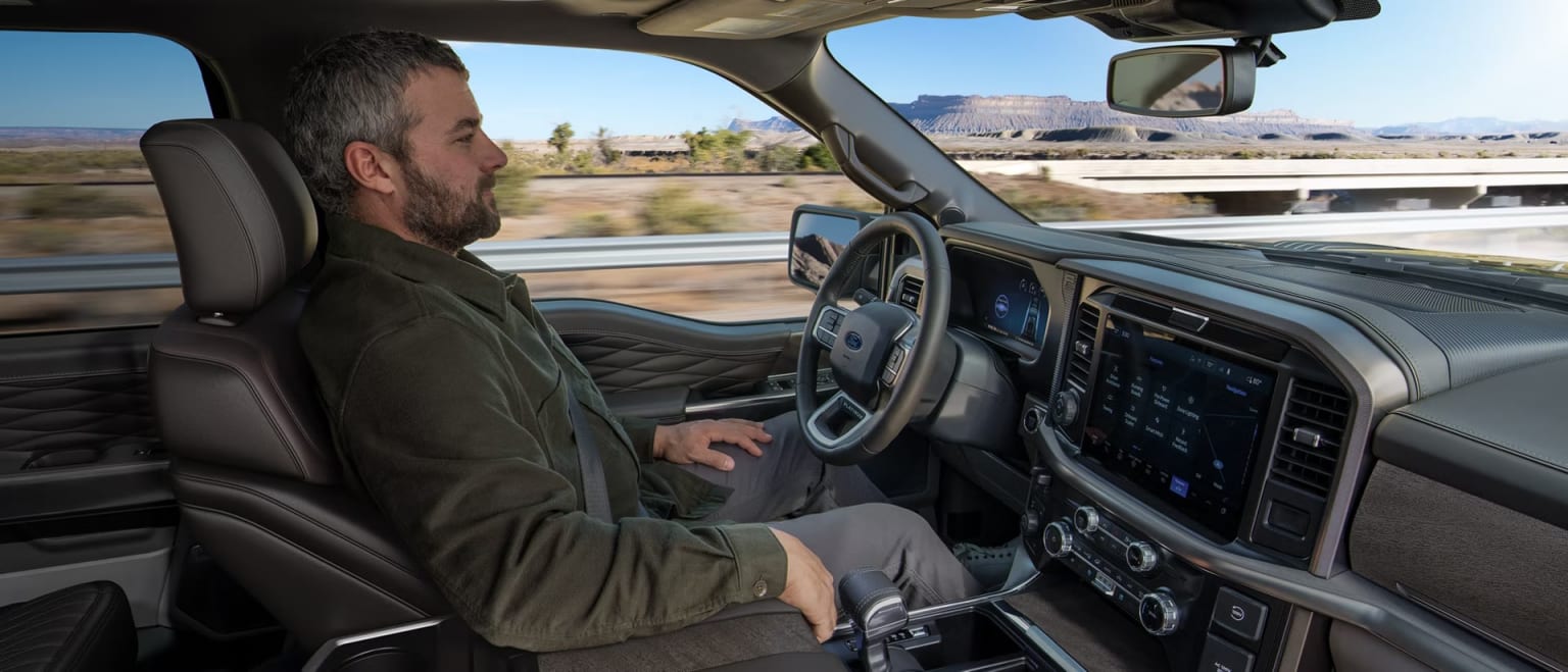 A man with a beard driving a modern truck through a desert landscape with mountains in the background.
