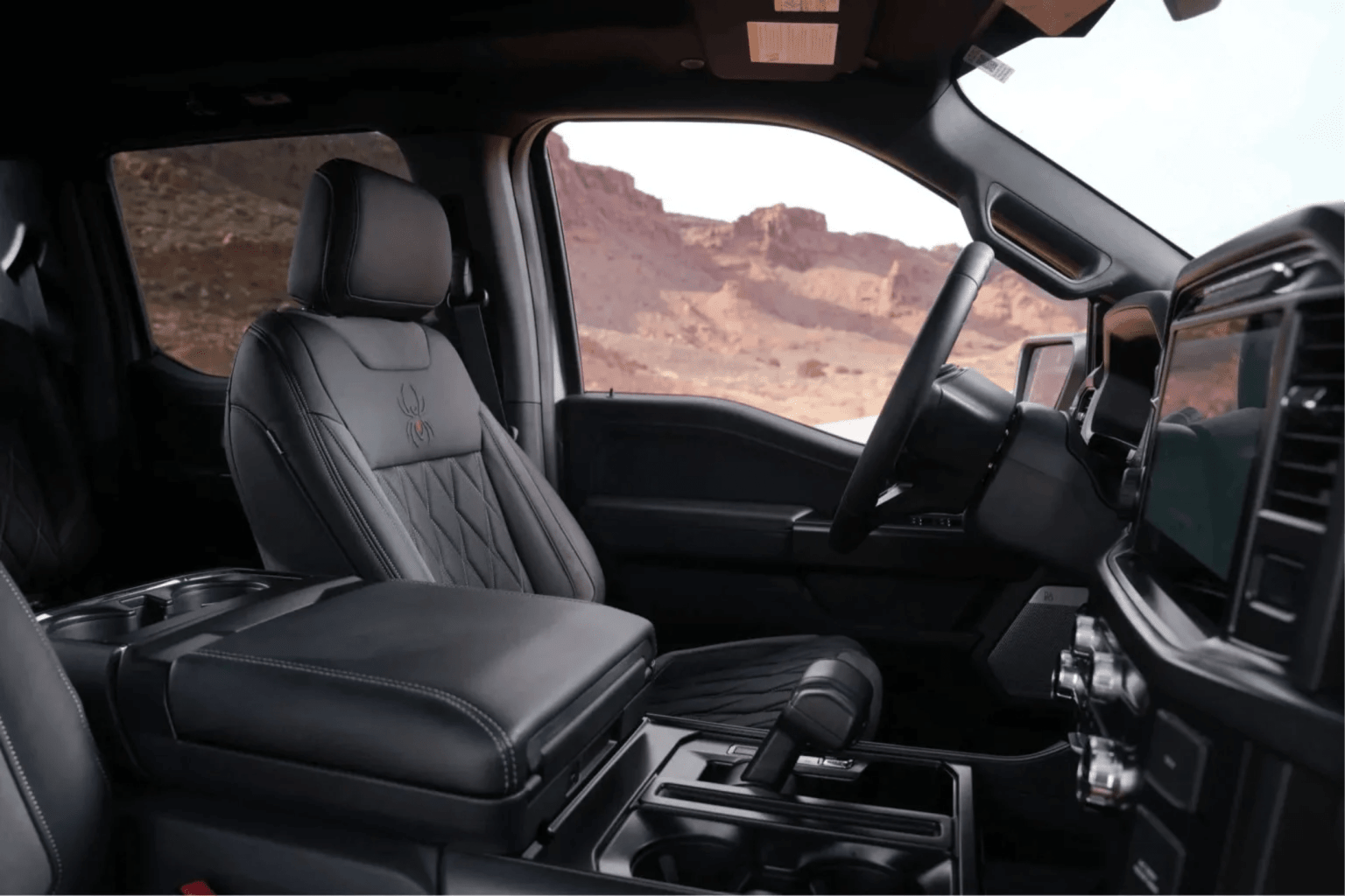 Interior of a black leather truck cabin with a quilted seat and spider emblem, with red rock formations visible through the windows.