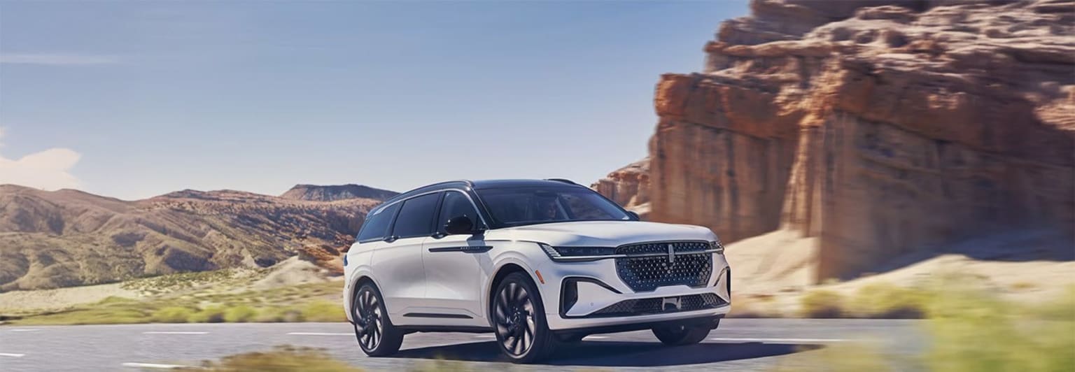 White luxury SUV driving on winding road through rugged mountainous landscape with clear blue sky in background.