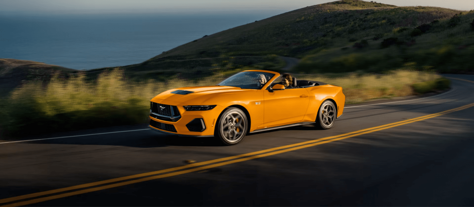 A bright orange convertible car drives along a winding coastal road with green hills and the ocean in the background.