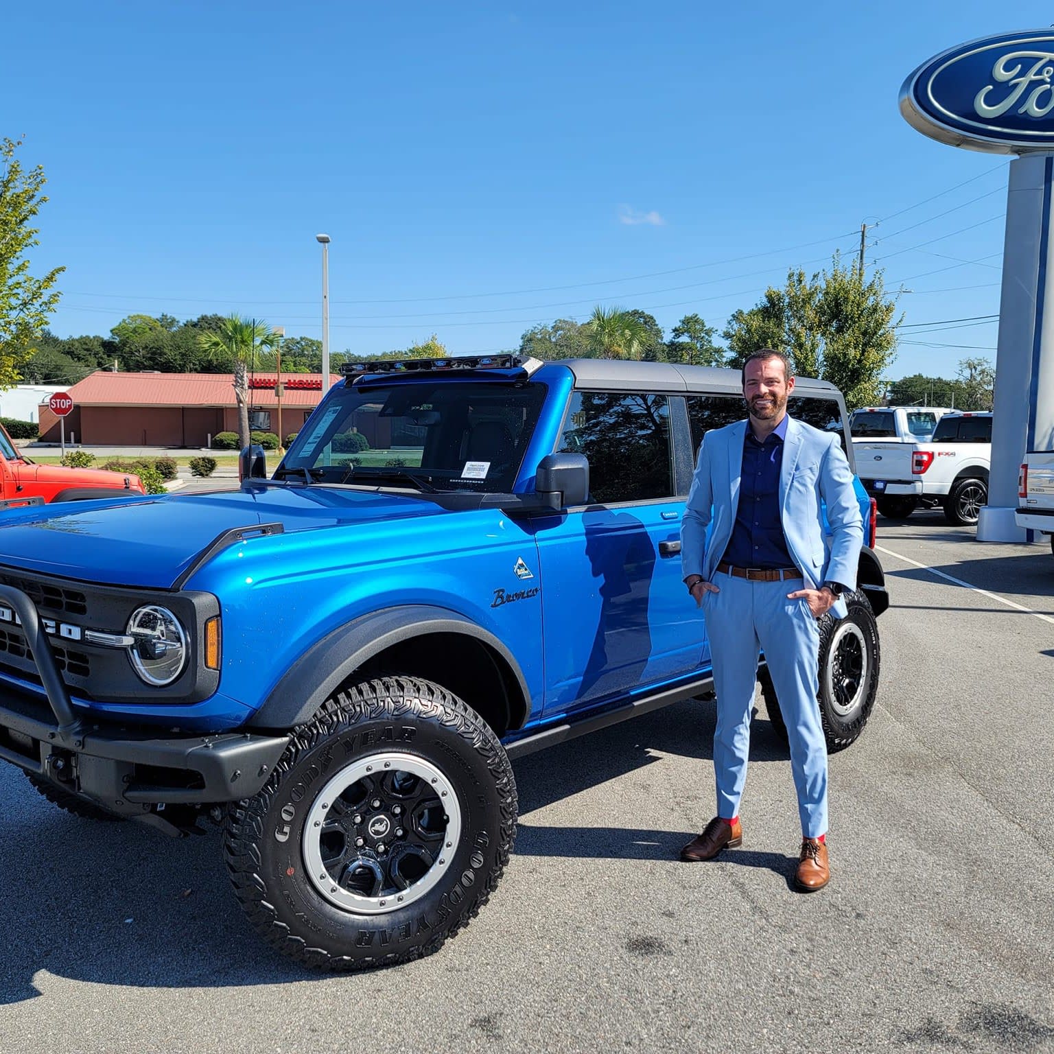A man stands next to a bright blue SUV in a car dealership lot, with a logo visible in the background.