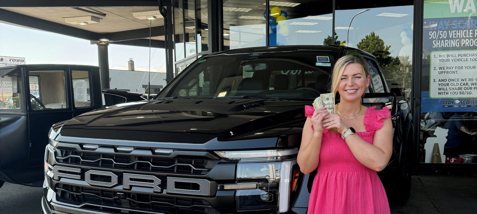A woman in a pink dress stands in front of a large black truck, with a car dealership in the background.