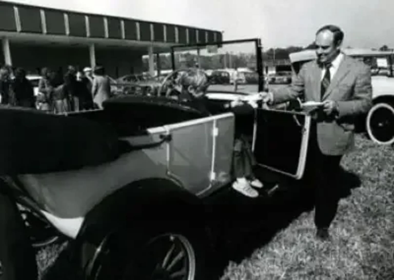 a man in a suit and tie standing next to an old car in front of a crowd of people