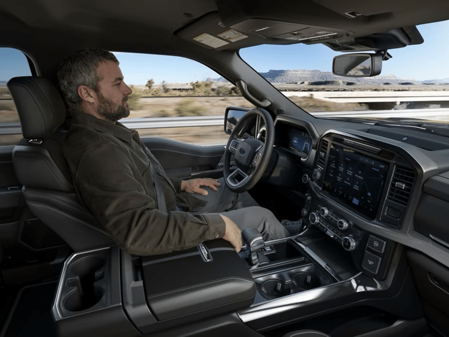 A person sitting in the driver's seat of a vehicle, with a dashboard and steering wheel visible in the foreground, and a landscape visible through the windshield in the background.