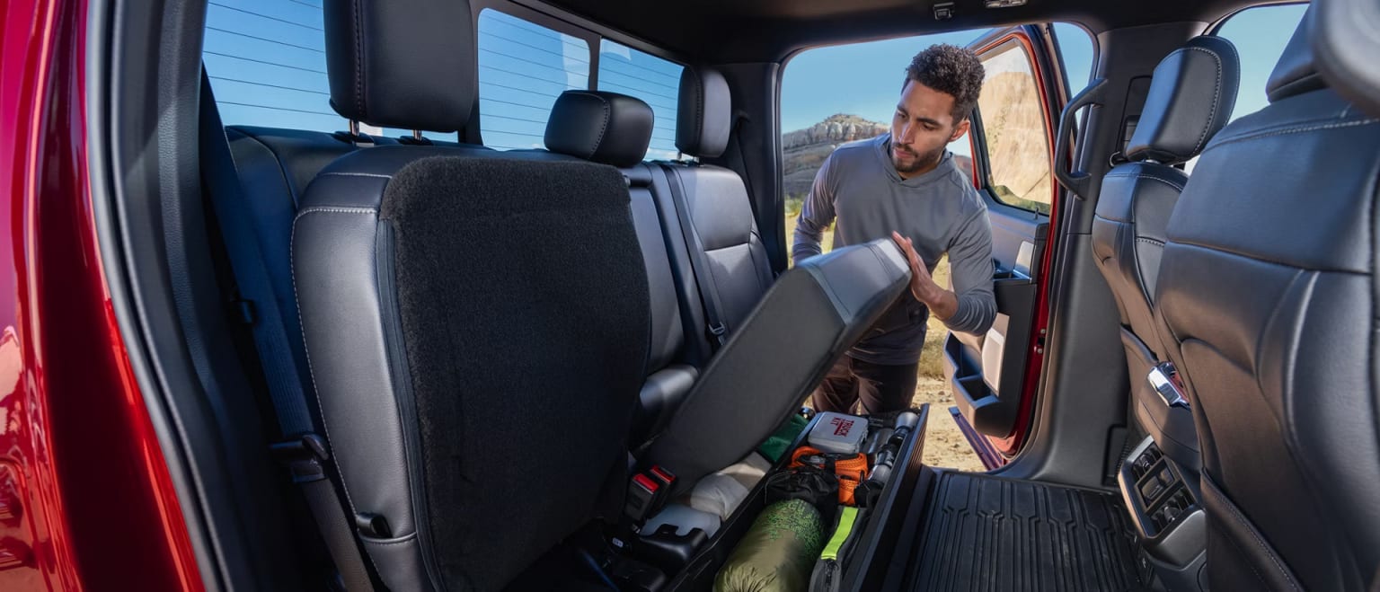 A man is organizing camping gear under the folded rear seats of a red truck with a desert landscape in the background.