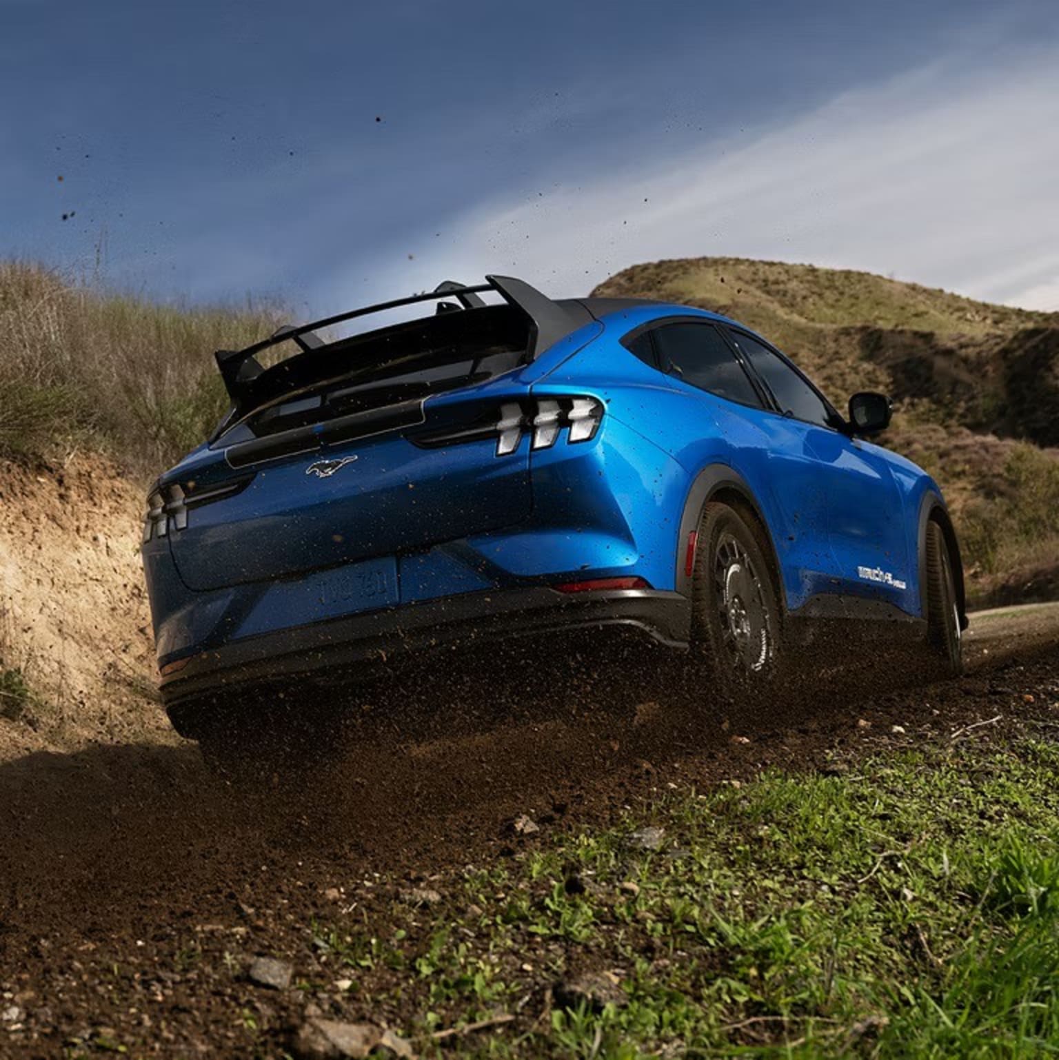 A blue sports car with a large rear spoiler driving through grassy, hilly terrain with a cloudy sky in the background.