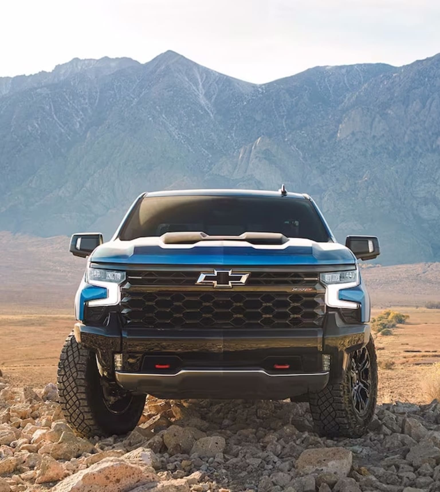 A blue pickup truck parked on a rocky hillside with mountains in the background, creating a rugged, remote atmosphere.