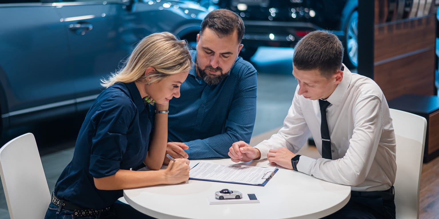 A couple signs paperwork at a table with a car salesman, while a blue SUV is visible in the background.