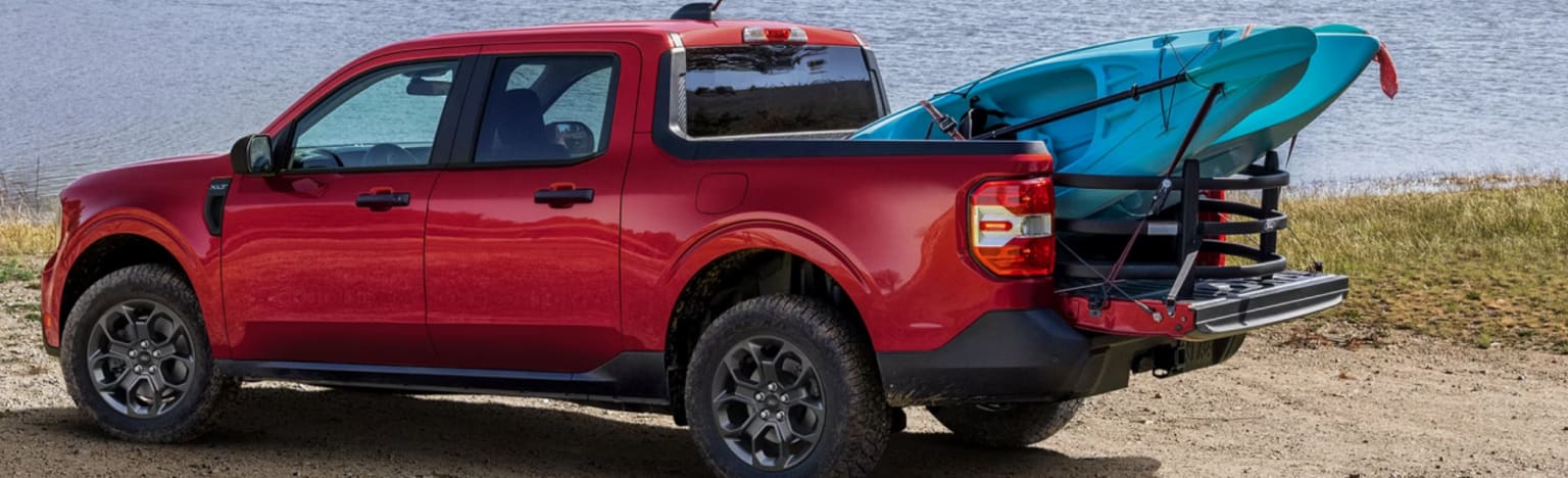 Red pickup truck with a kayak in the bed parked on a sandy shore with a body of water in the background.