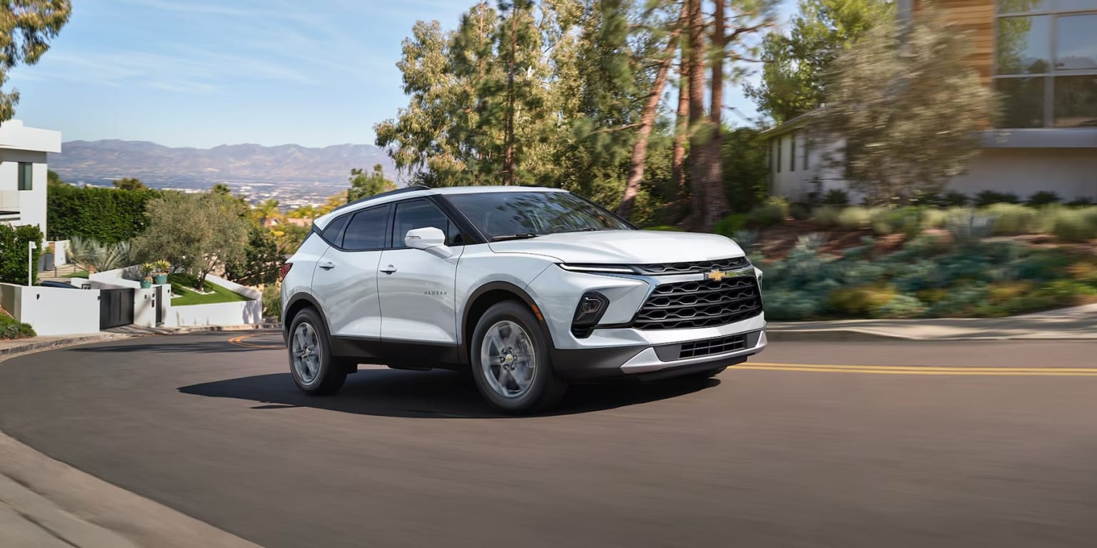 A white SUV is driving down a street, with a house in the background.