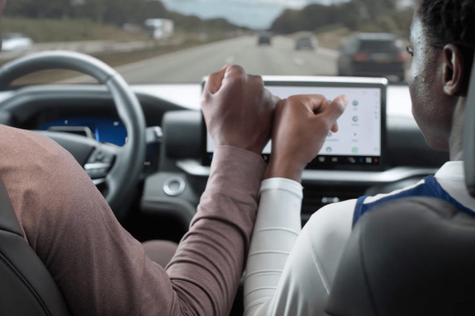 Two people hold hands in the foreground while driving on a highway in the background.