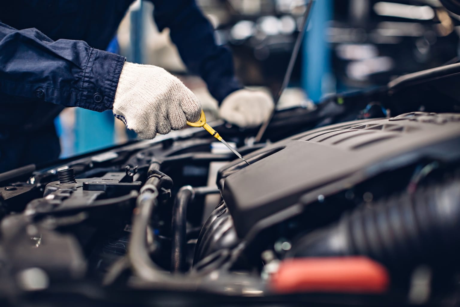 A mechanic in gloves checks the oil level of a car engine inside a blurred garage setting.