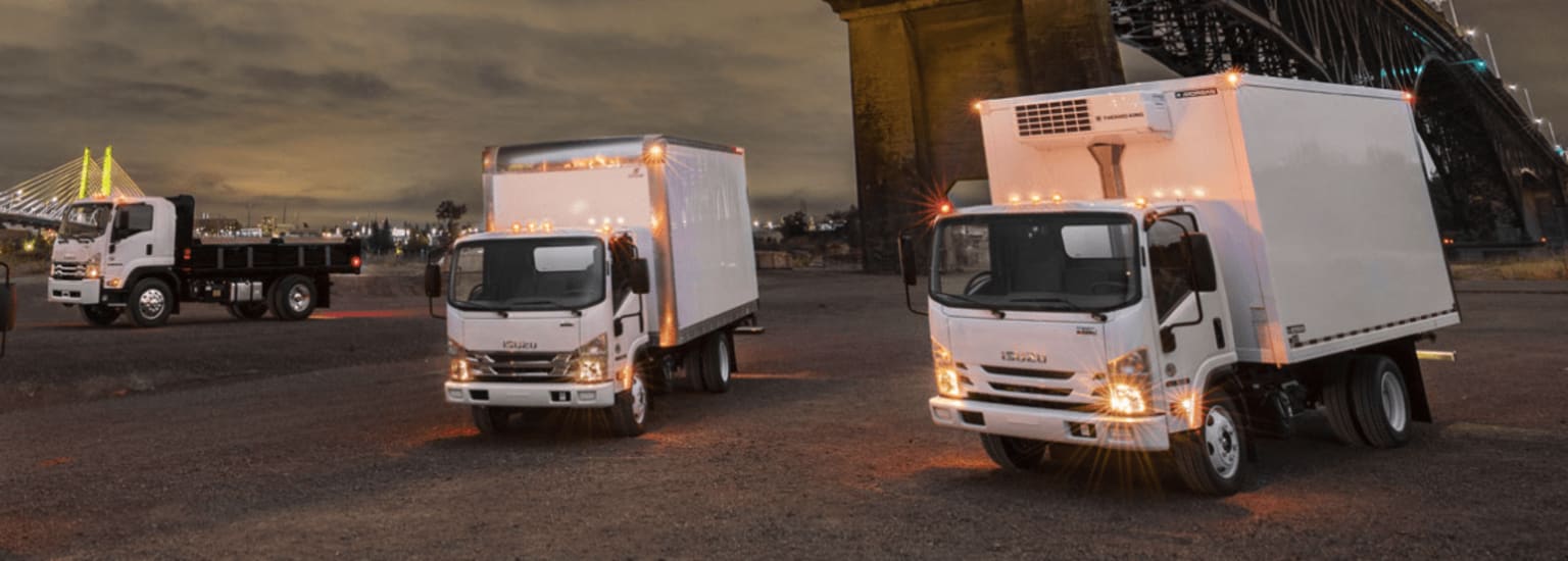 Two white refrigerated delivery trucks parked on a dimly lit street, with a large building or structure visible in the background.