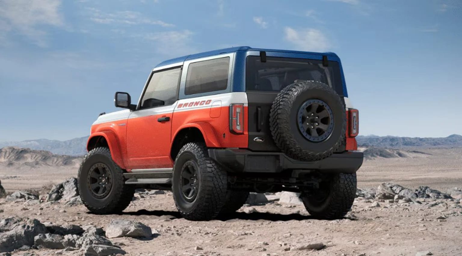 An off-road vehicle with large tires in orange and blue colors drives through a rocky desert landscape with mountains in the background.