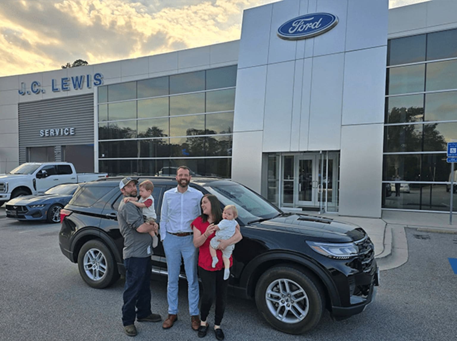 A family of four, including two adults and two children, standing in front of a dealership with a large logo visible on the building.