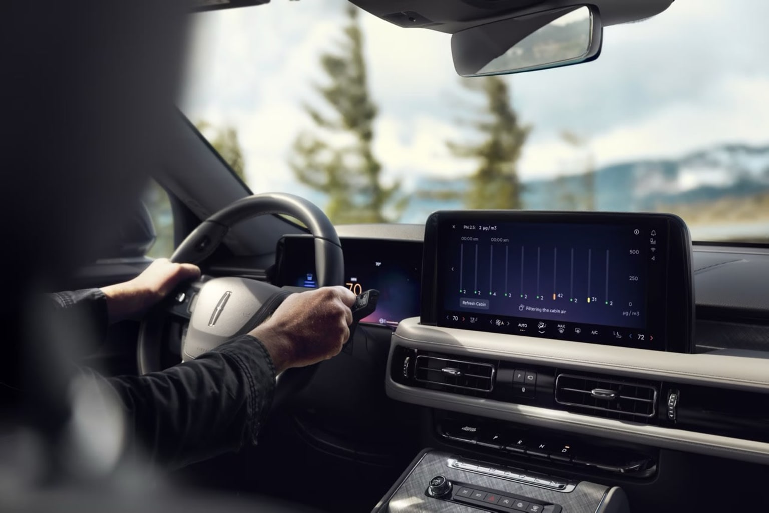 Interior view of a car with driver's hands on the steering wheel, touchscreen display in the center console, and scenic forested background outside.