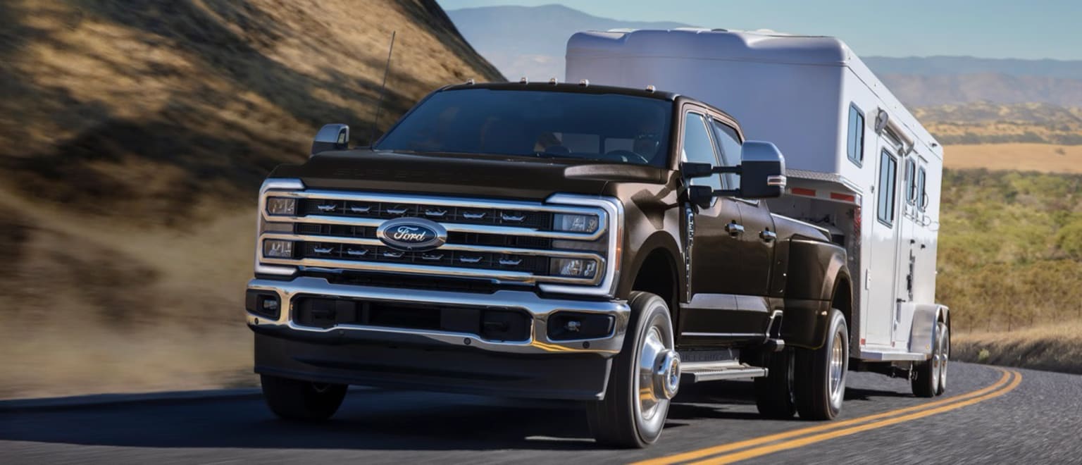 A large black pickup truck towing a white recreational vehicle on a winding mountain road, with rugged terrain and mountains visible in the background.