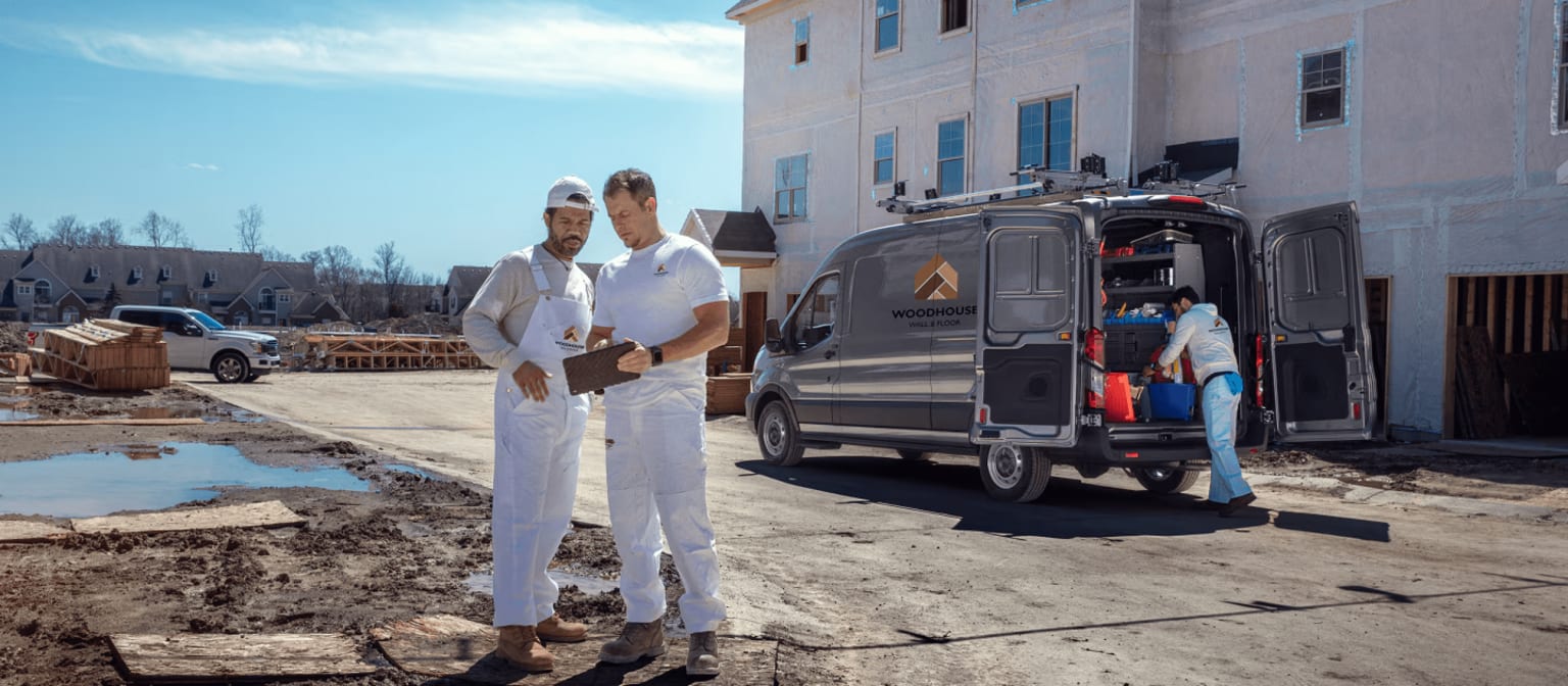 Two painters in white overalls stand on a muddy construction site, reviewing a tablet, while a van is being loaded with supplies in front of a building under construction.