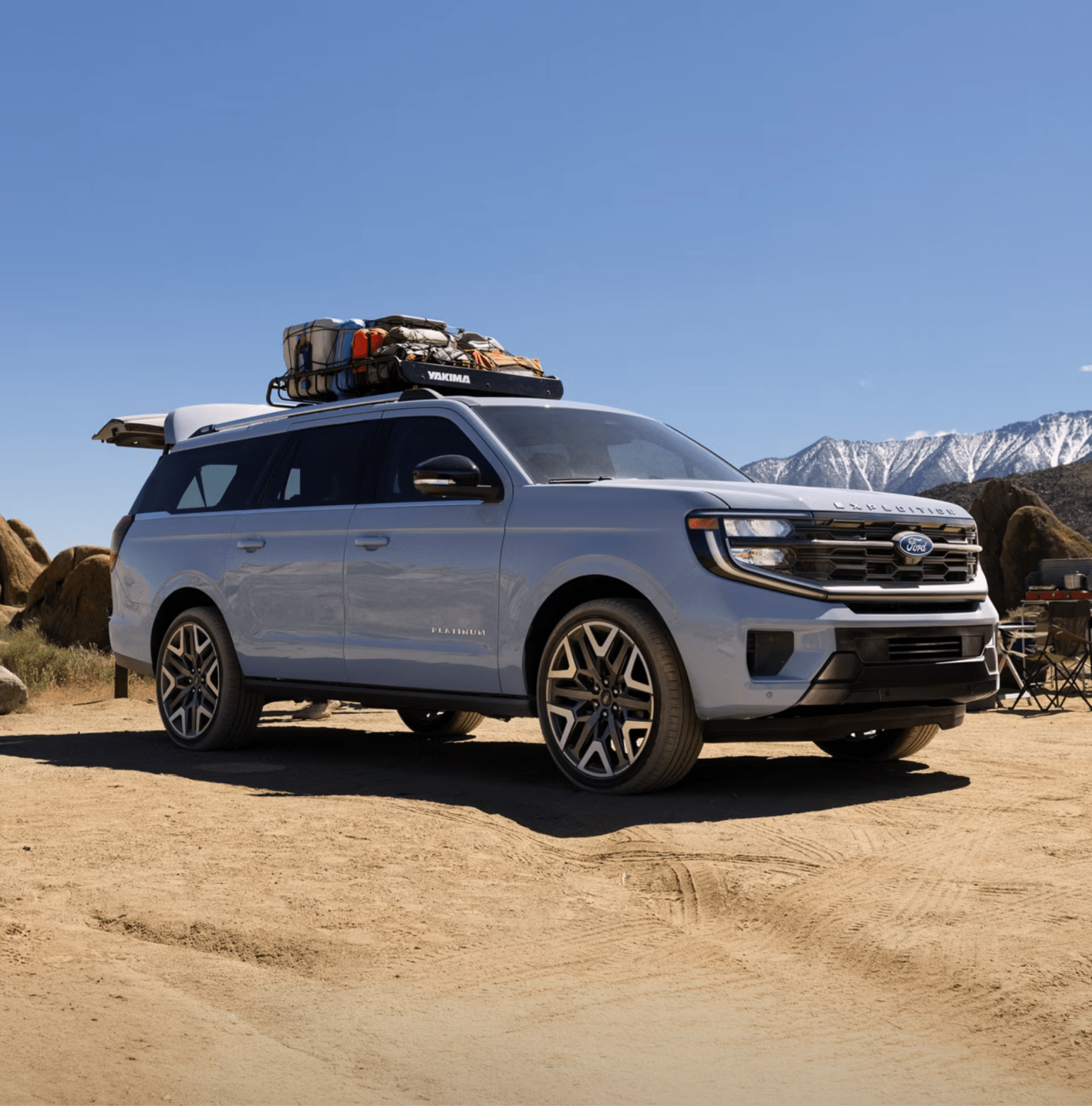 A light blue SUV with a roof rack loaded with gear on a dirt road, with rocky desert terrain and snow-capped mountains in the background.