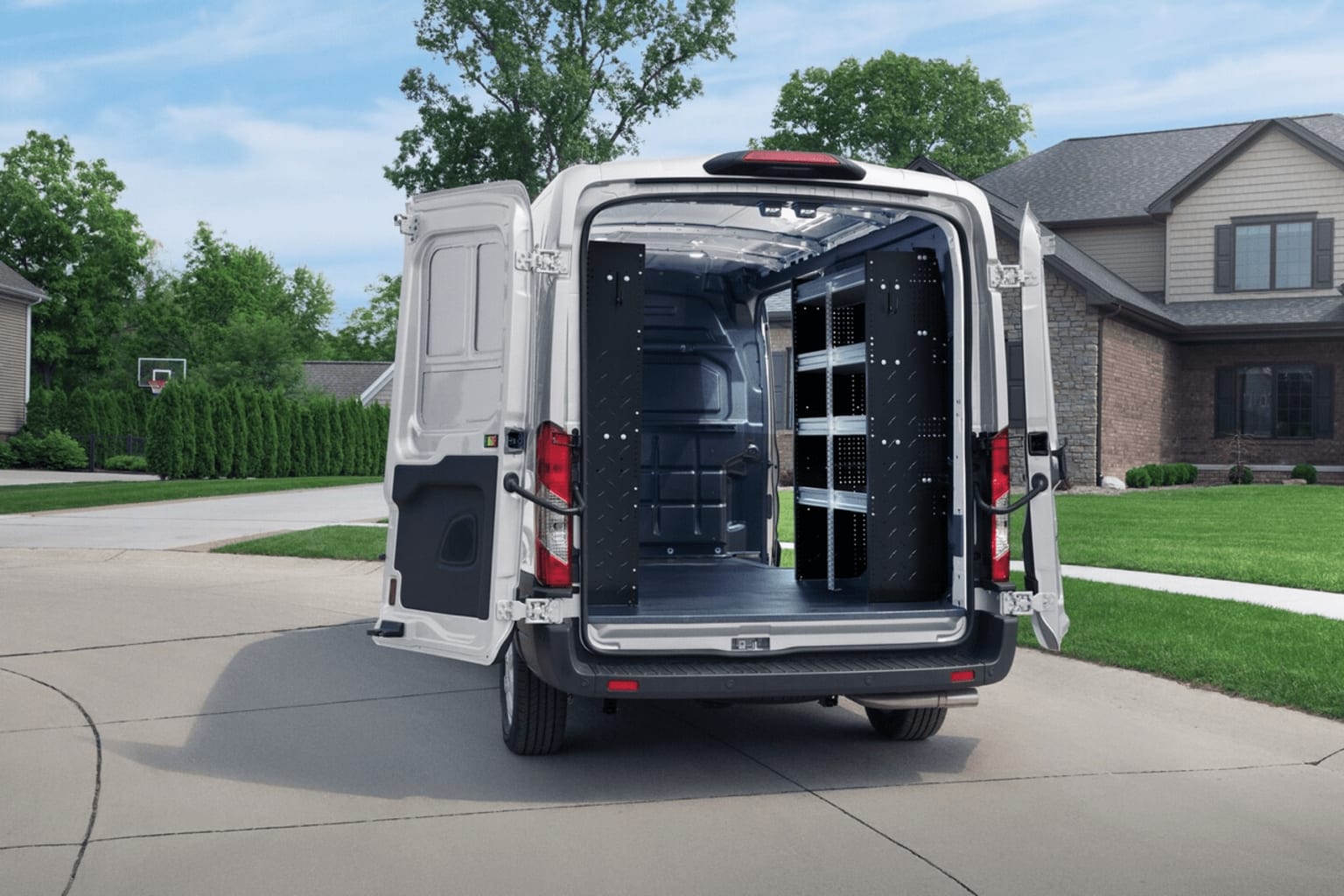 A white cargo van with shelves and storage units visible in the open rear compartment, parked on a paved driveway in front of a residential neighborhood with trees and houses in the background.