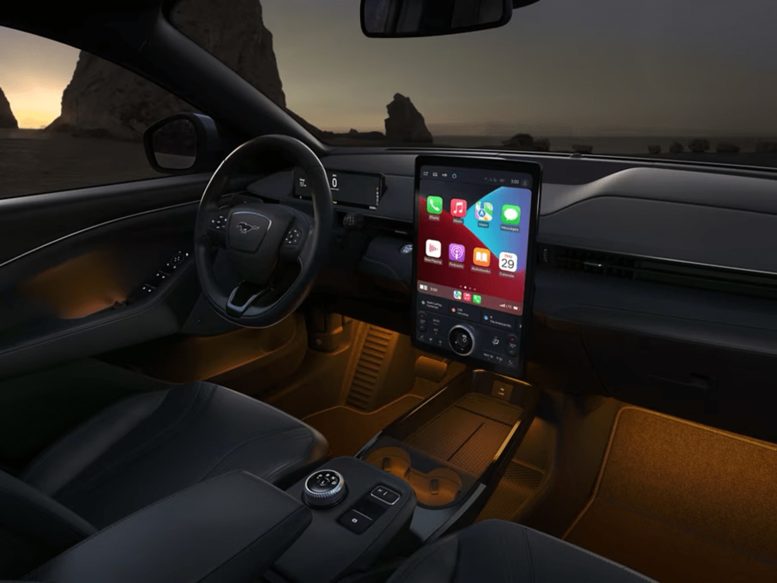 Interior of a vehicle at sunset with a large touchscreen display and various controls visible in the foreground, while a mountainous landscape can be seen through the windshield in the background.