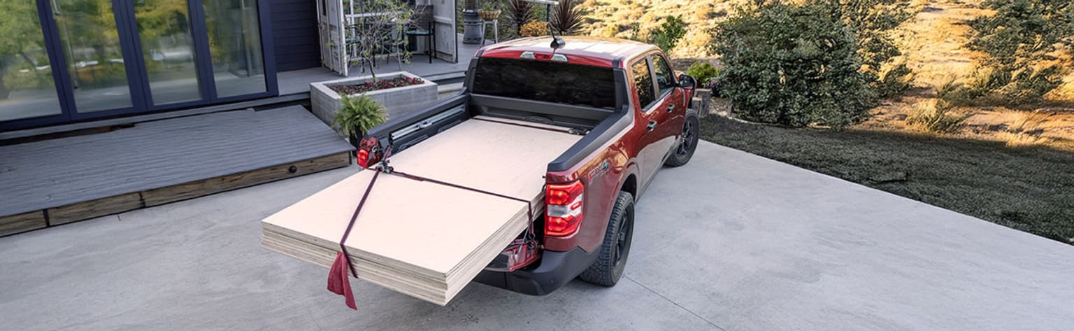 A red pickup truck with a cargo bed full of stacked boxes is parked on a concrete patio, surrounded by lush greenery in the background.