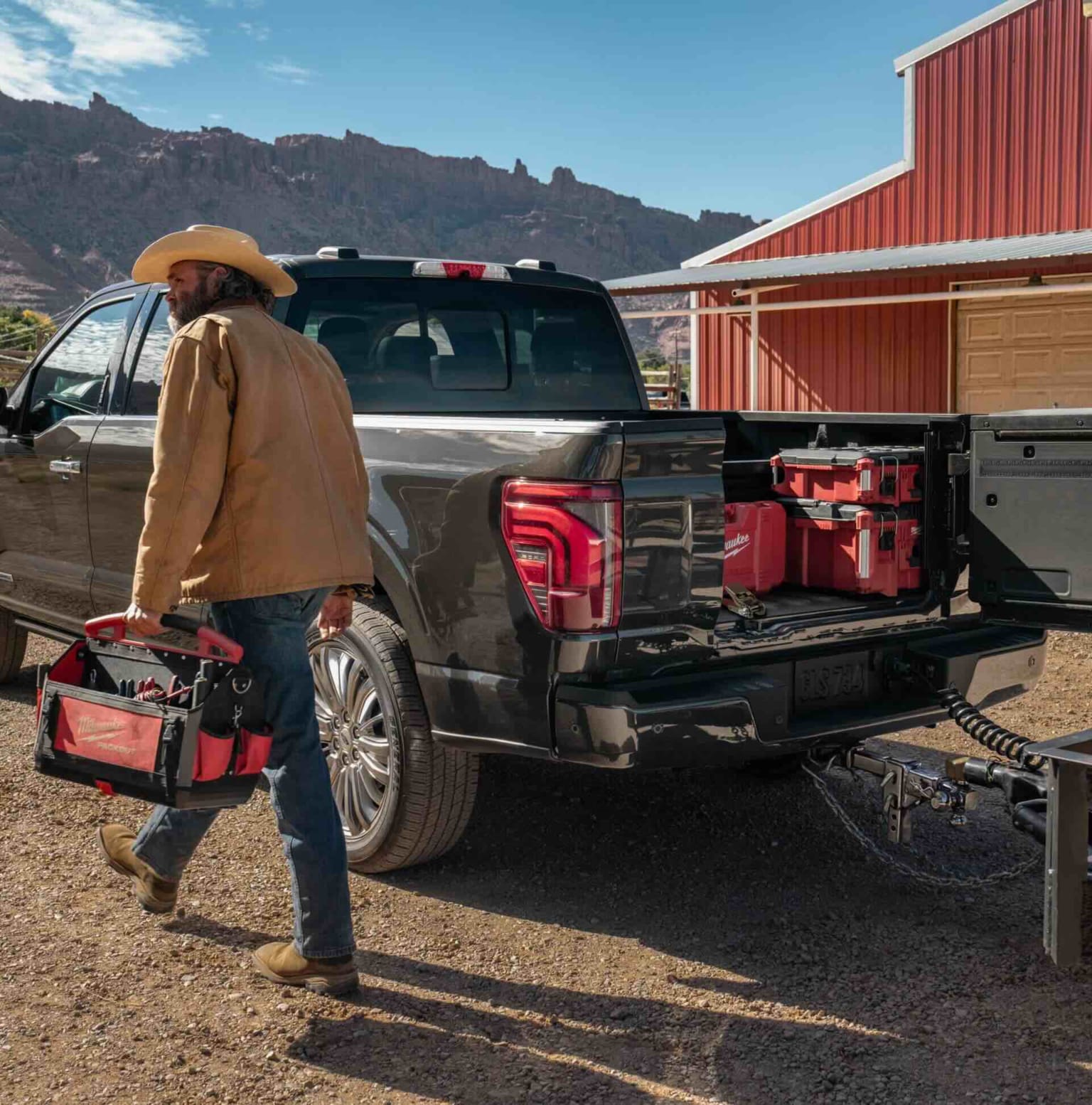 A man in a cowboy hat carrying a red toolbox walking towards a dark pickup truck with red toolboxes in its open bed, with a red barn in the background.