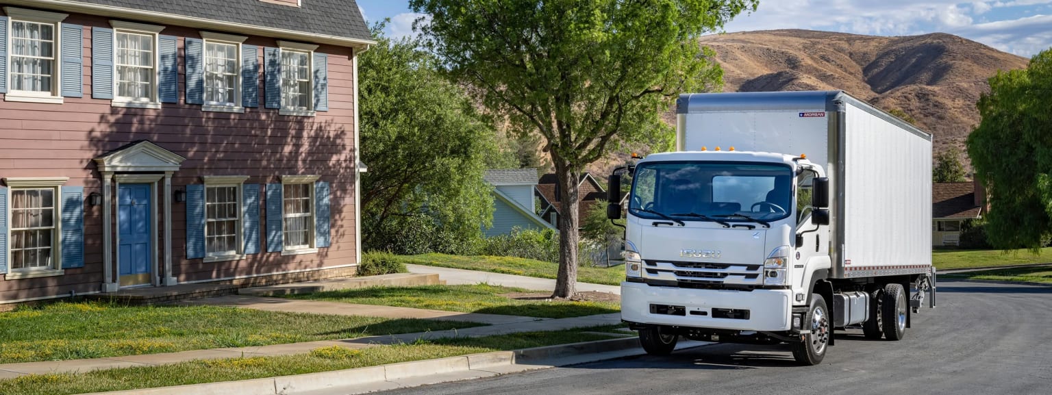 A large white delivery truck parked on a residential street surrounded by colorful houses and lush trees.