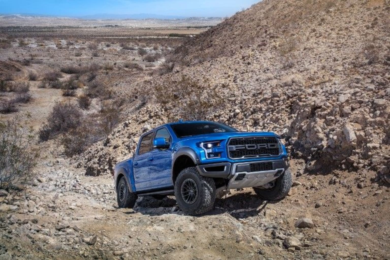 a blue truck is driving on a dirt road in the desert with a hill in the background and a blue sky in the background