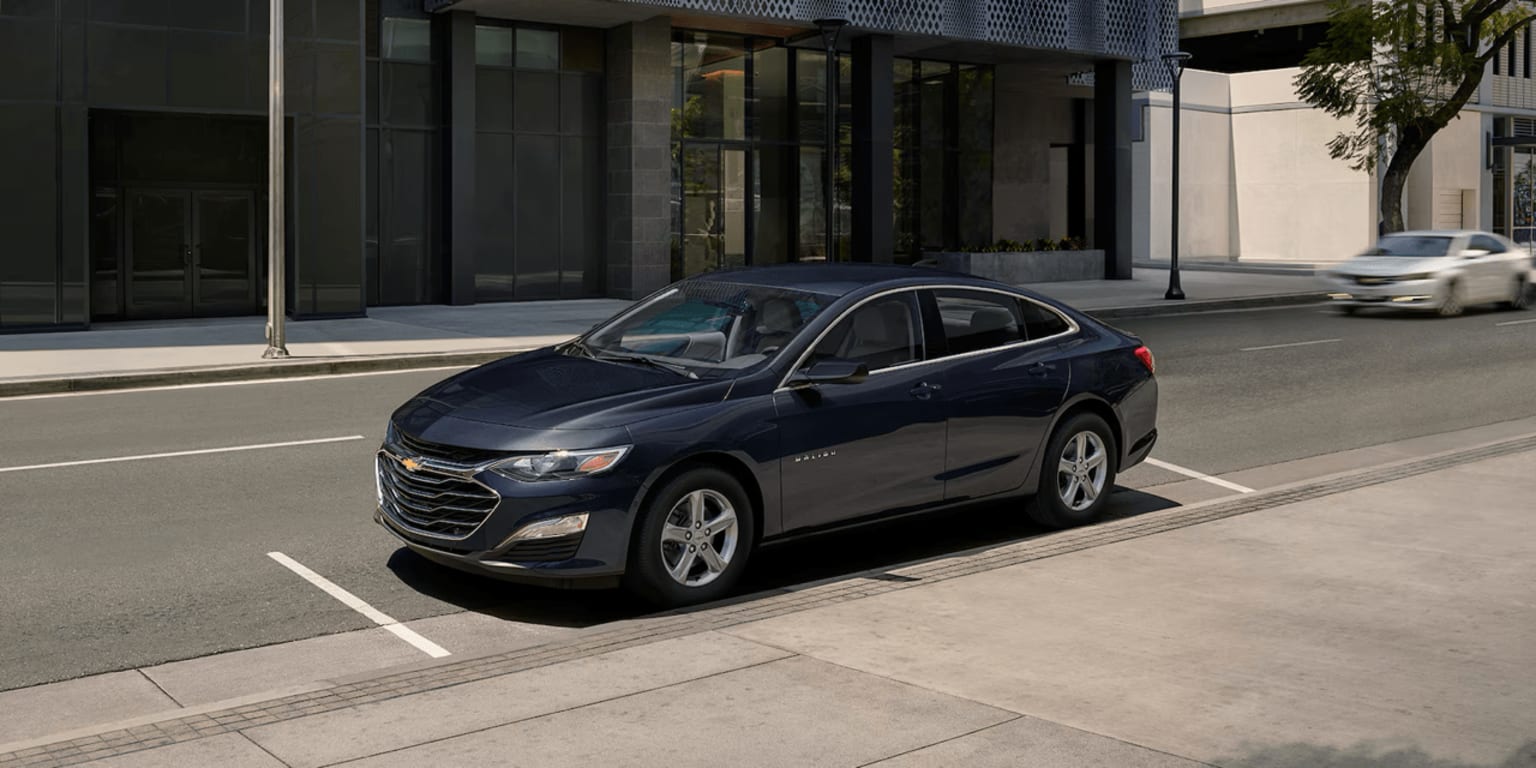 A dark blue SUV parked on the street in front of a modern building, with other vehicles passing by in the background.