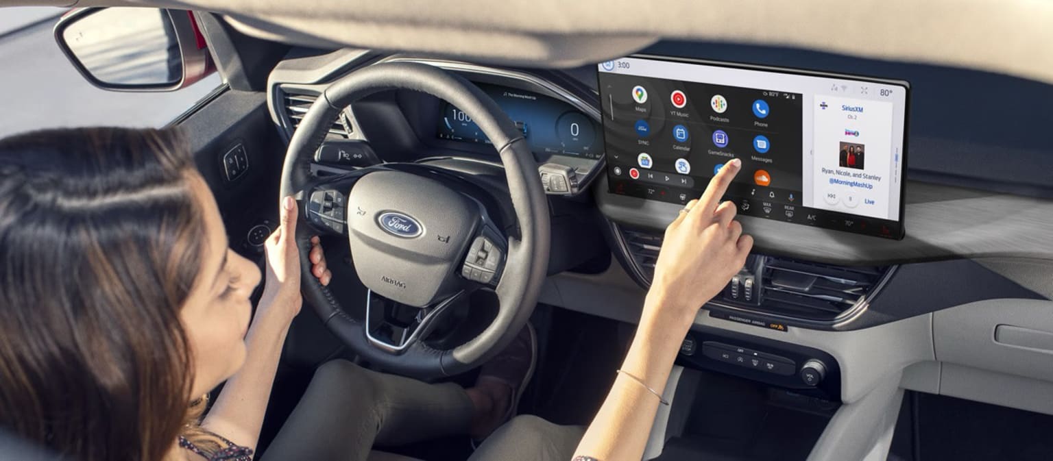 The image displays the interior of a contemporary vehicle, featuring a person's hands on the steering wheel and a prominent touchscreen display in the center console.