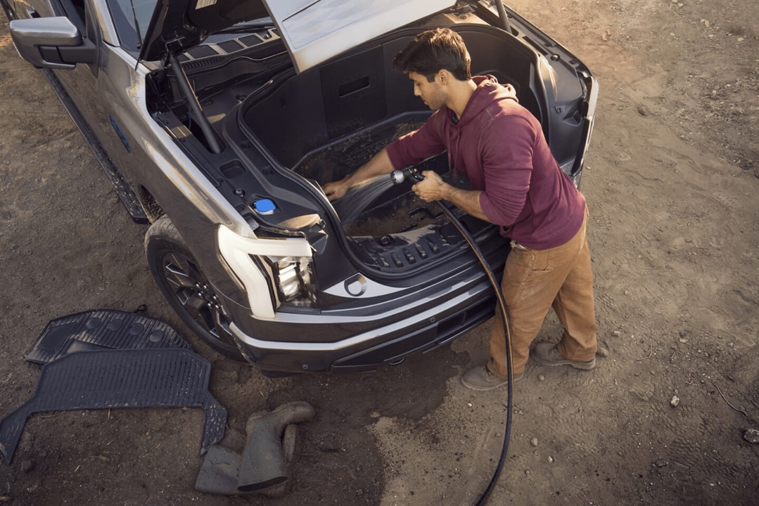 A person in a shirt is working on the open trunk of a dark-colored vehicle, surrounded by various tools and equipment on the ground.