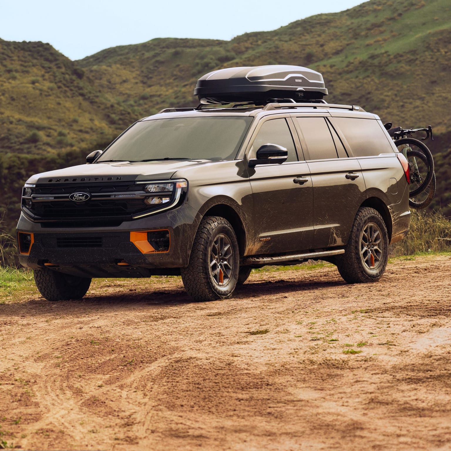 An off-road SUV with roof rack and bike rack on a dirt road surrounded by hilly grassy terrain.