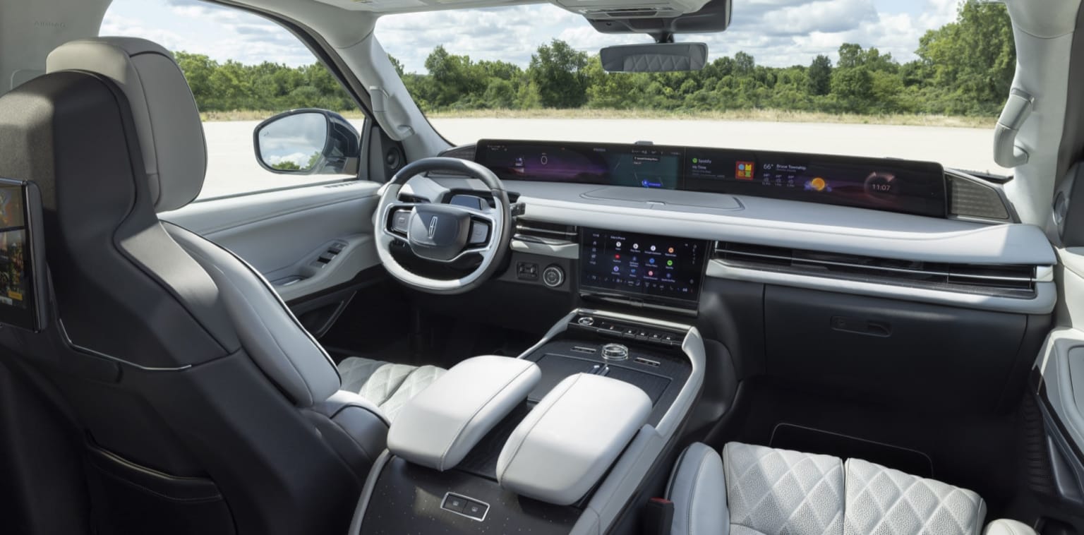 The image shows the interior of a modern, high-tech vehicle with a sleek dashboard, a large central display, and a steering wheel. The background outside the vehicle appears to be a rural or forested area.
