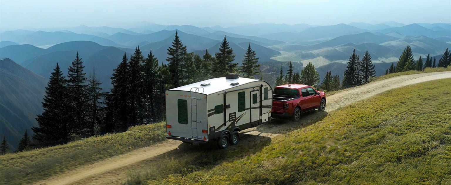 A small white camper trailer being towed by a red pickup truck on a winding dirt road, surrounded by lush green hills and towering pine trees in the distance.