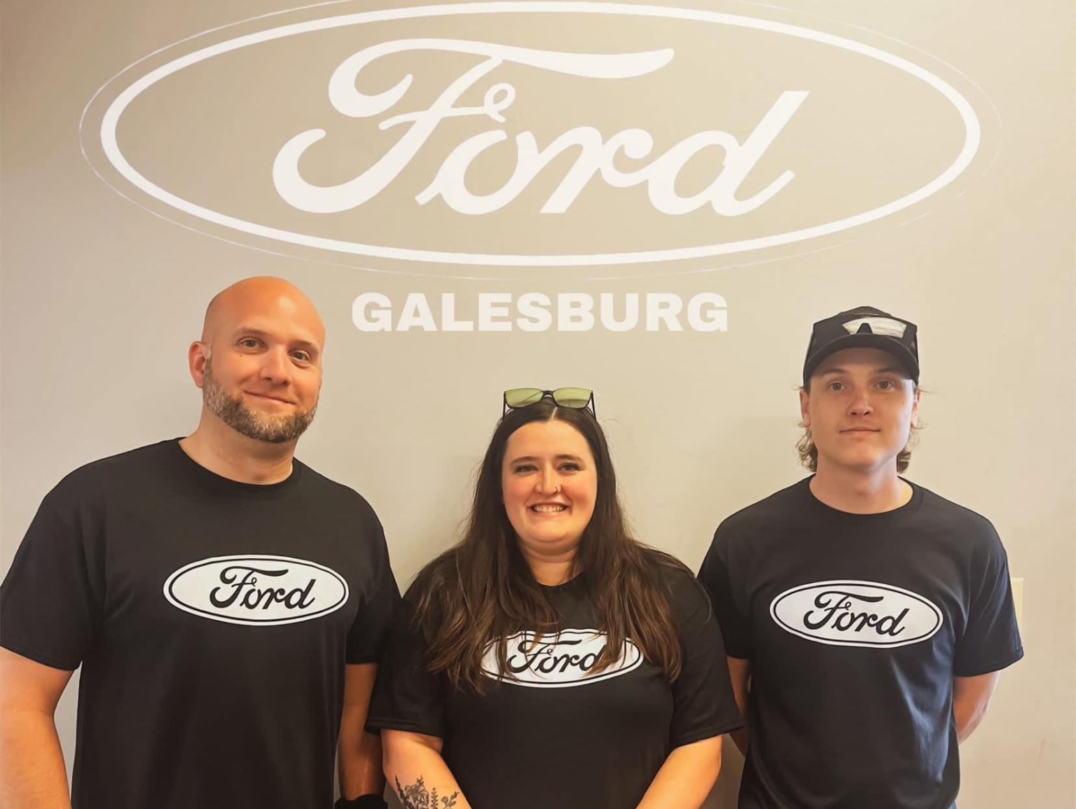 The image shows three people wearing company-branded t-shirts standing in front of a dealership sign in Galesburg.