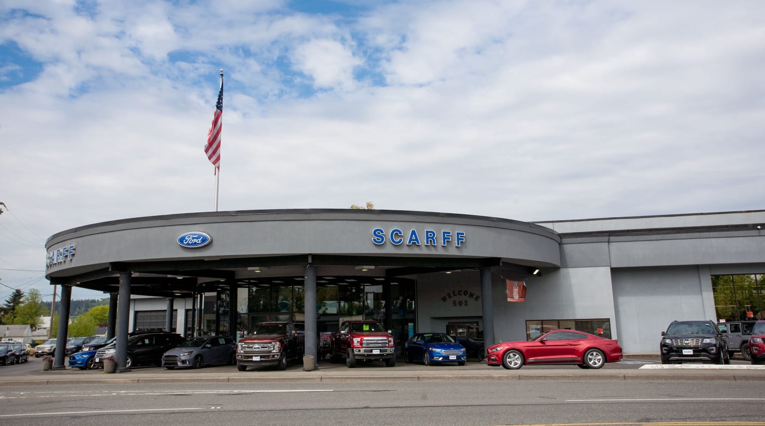 A dealership with several vehicles parked in front, set against a cloudy blue sky.