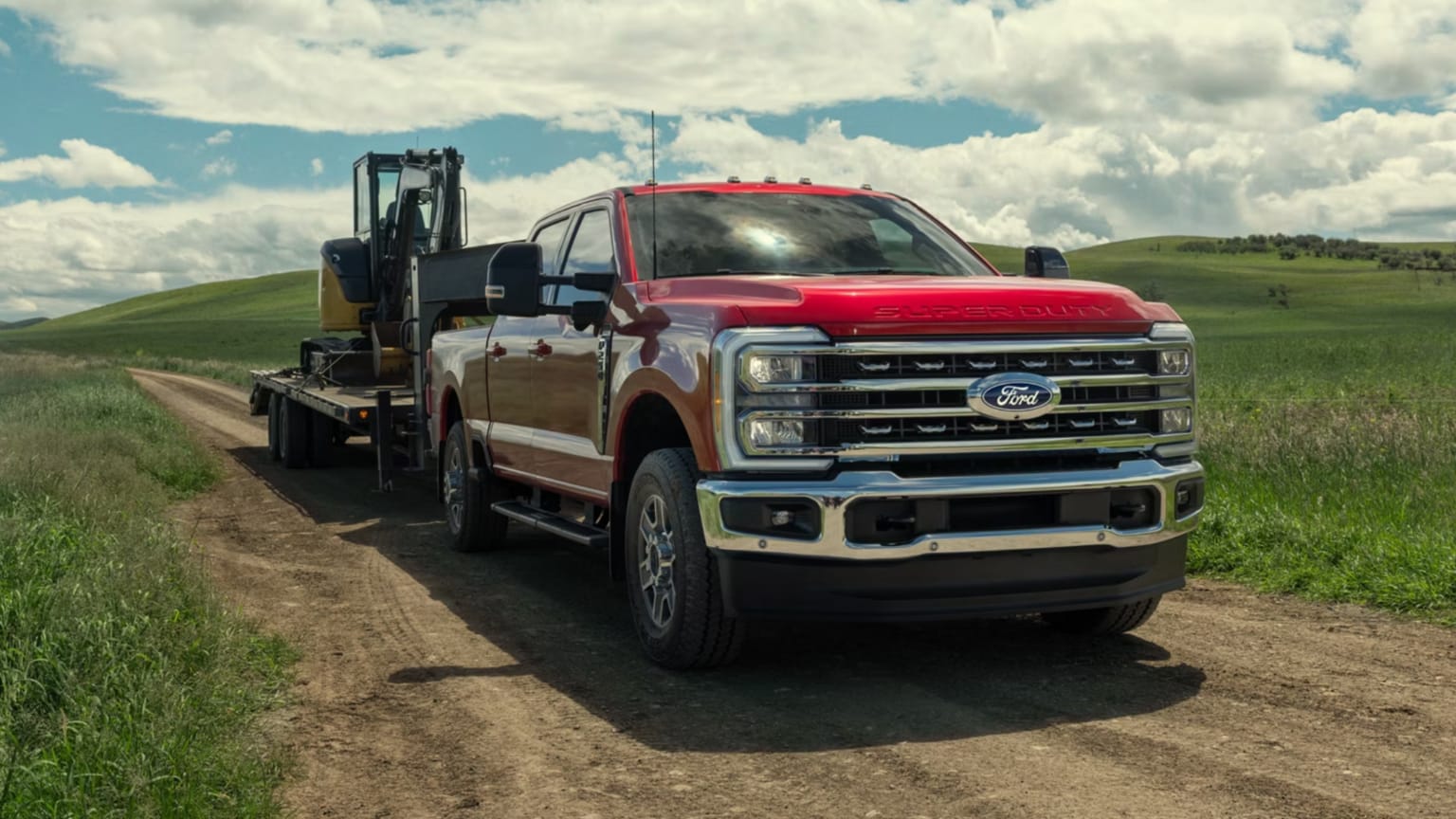 Red pickup truck towing a trailer on a dirt road amidst green hills under a cloudy sky.
