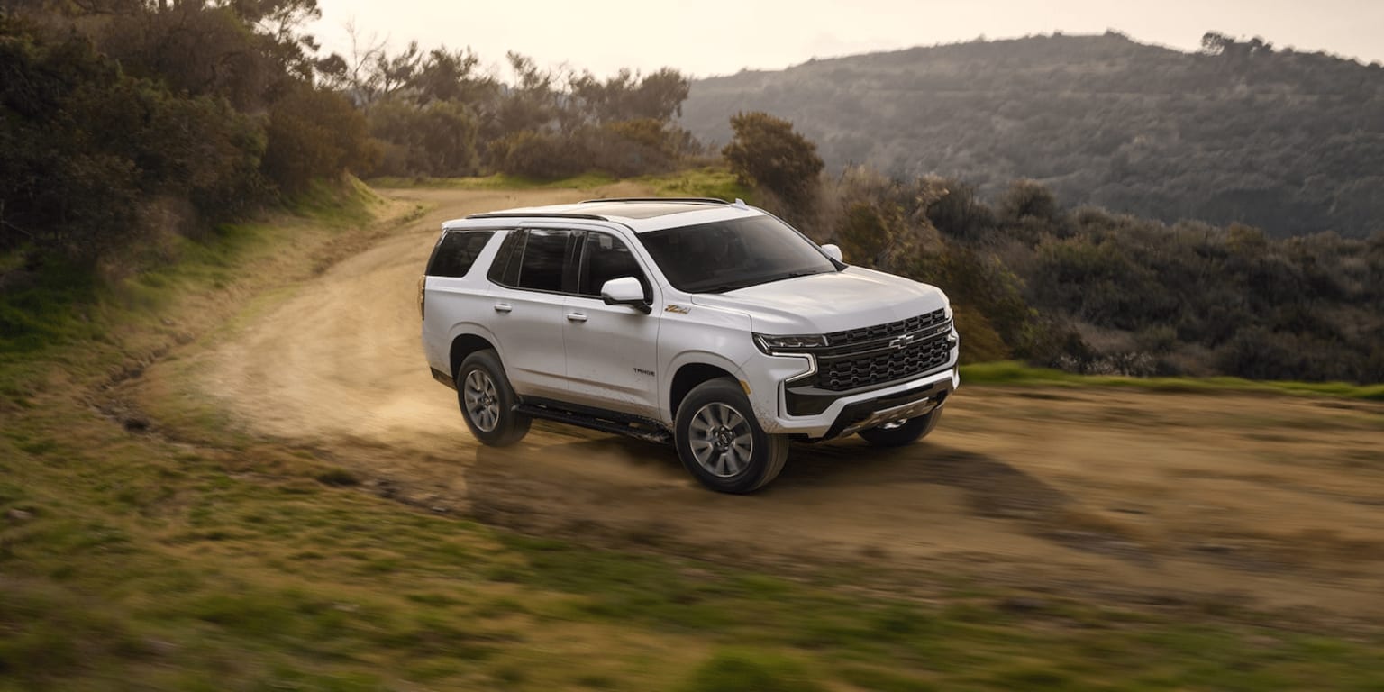 The image shows a white SUV driving on a dirt road through a lush, forested landscape with hills and mountains in the background.