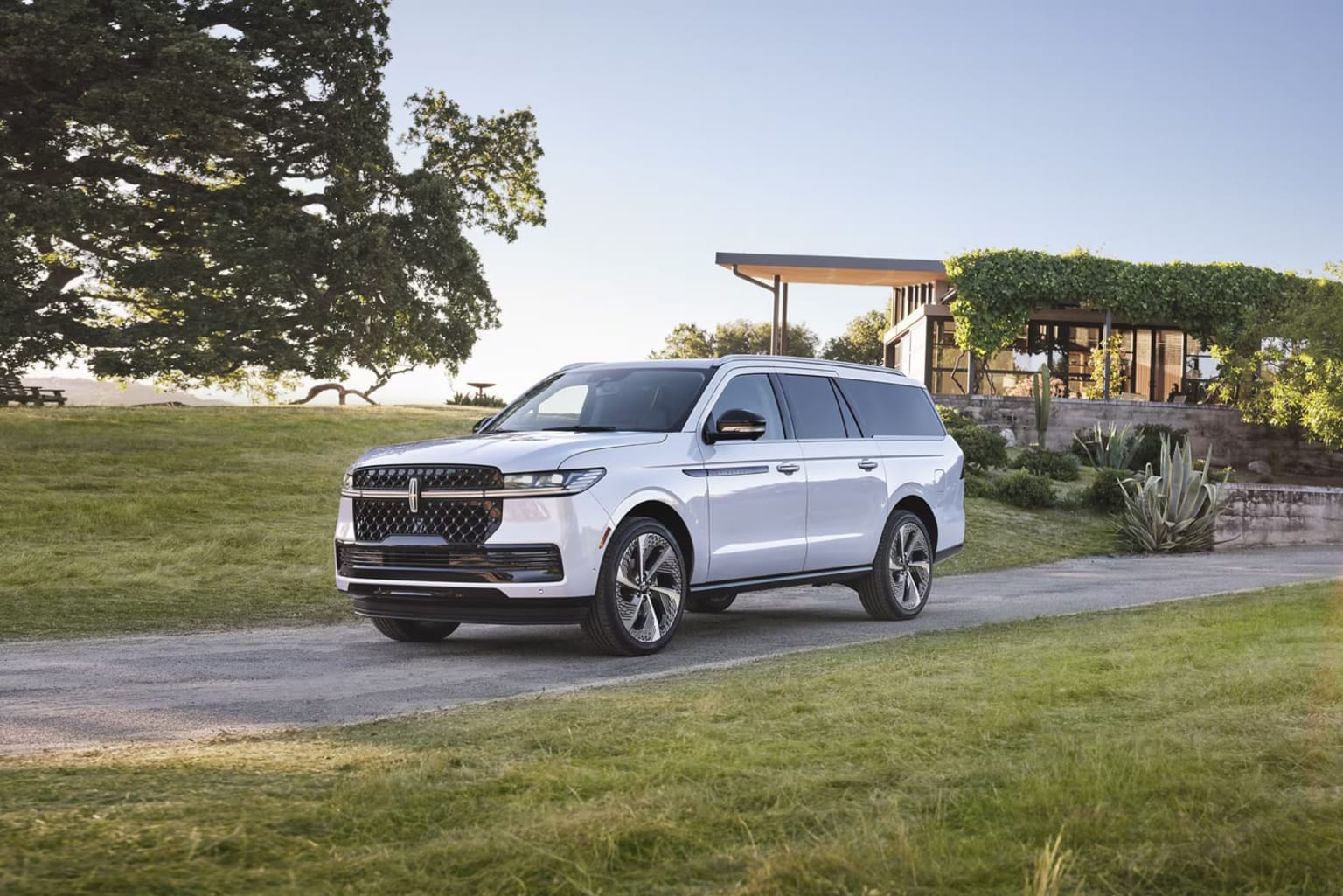 Large white SUV parked on grassy field with wooden structure and lush greenery in background.