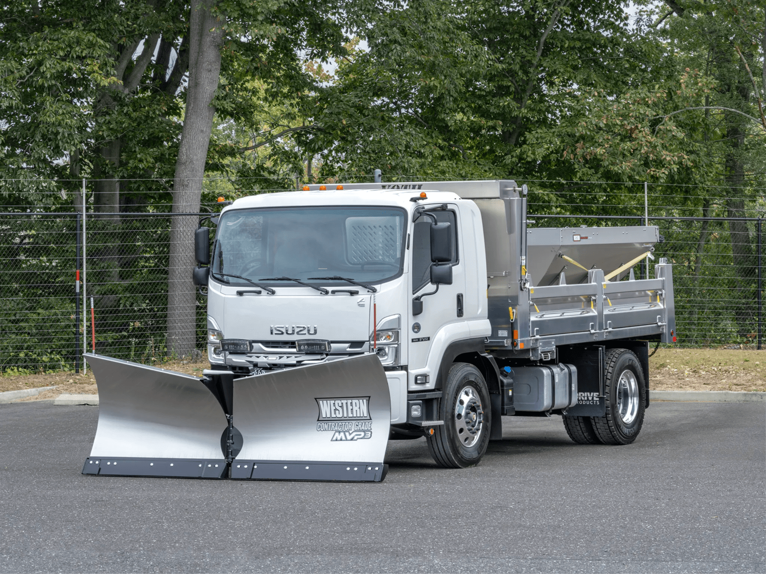 A white truck with a snowplow attachment on a paved surface in front of a chain-link fence and lush green trees.